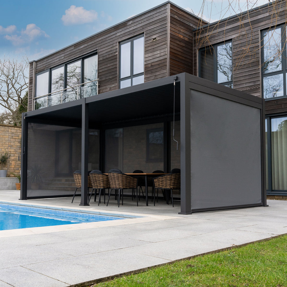 A modern outdoor patio pergola in Scotland with a built-in seating area, set in a lush green backyard. The pergola features a dark grey frame, a slatted roof with warm LED lighting, and a retractable side awning.